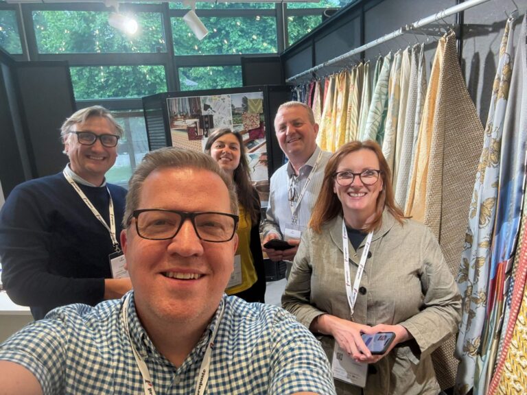 David Walters sales team at Proposte fair posing for a selfie in front of their stand with fabric hanging behind them.